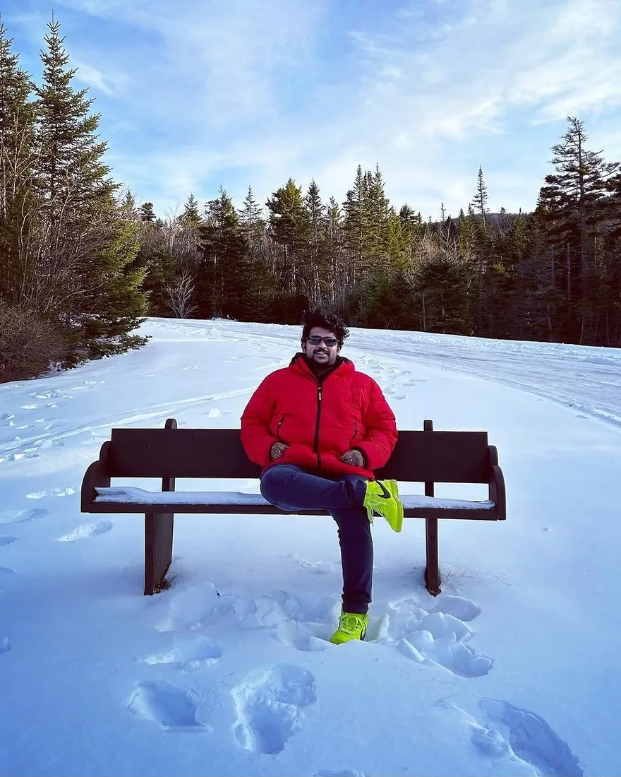 Sudhanshu in a red puffer jacket and sunglasses, sitting on a wooden bench in a snowy clearing ringed by evergreen trees under a blue sky.