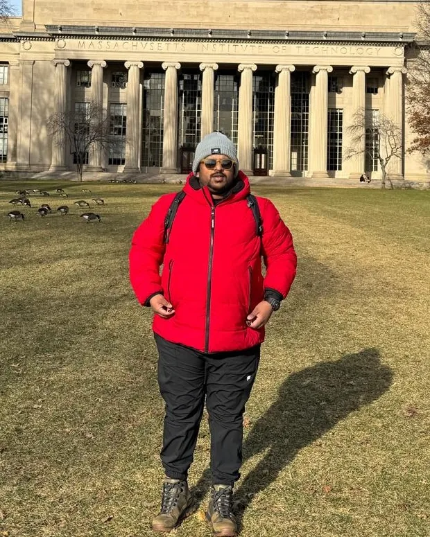Sudhanshu in a red winter jacket and grey beanie, standing on a lawn in front of a columned stone building.